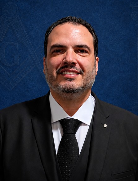Homem sorridente de terno escuro e gravata preta, fundo azul com padrão sutil, retrato próximo.